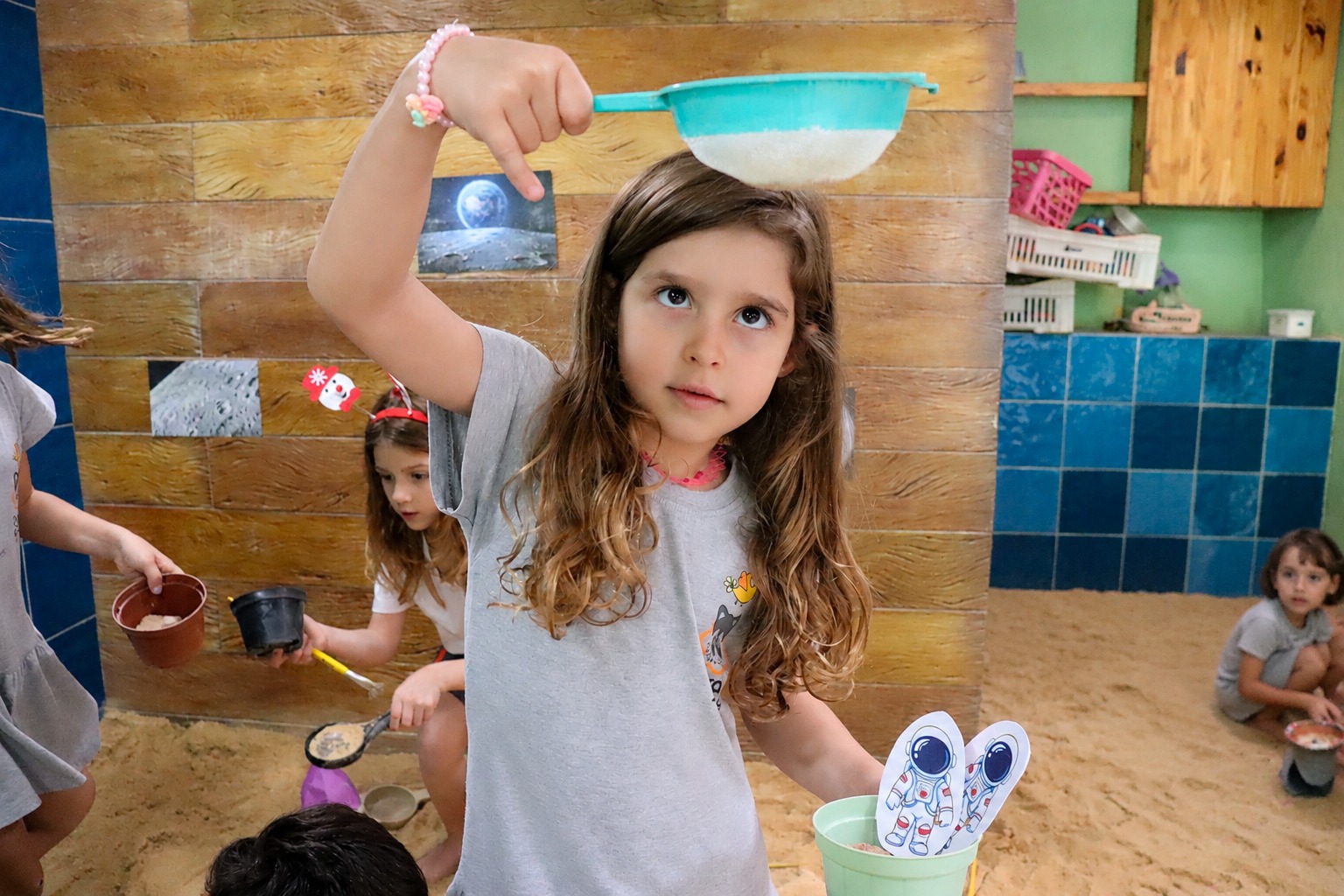 Menina peneirando areia com uma peneira azul na área de areia da escola, com colegas explorando materiais ao fundo