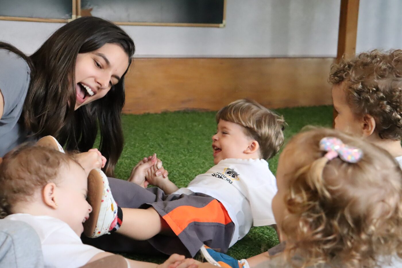 Educadora brincando no chão com crianças pequenas, todos rindo juntos