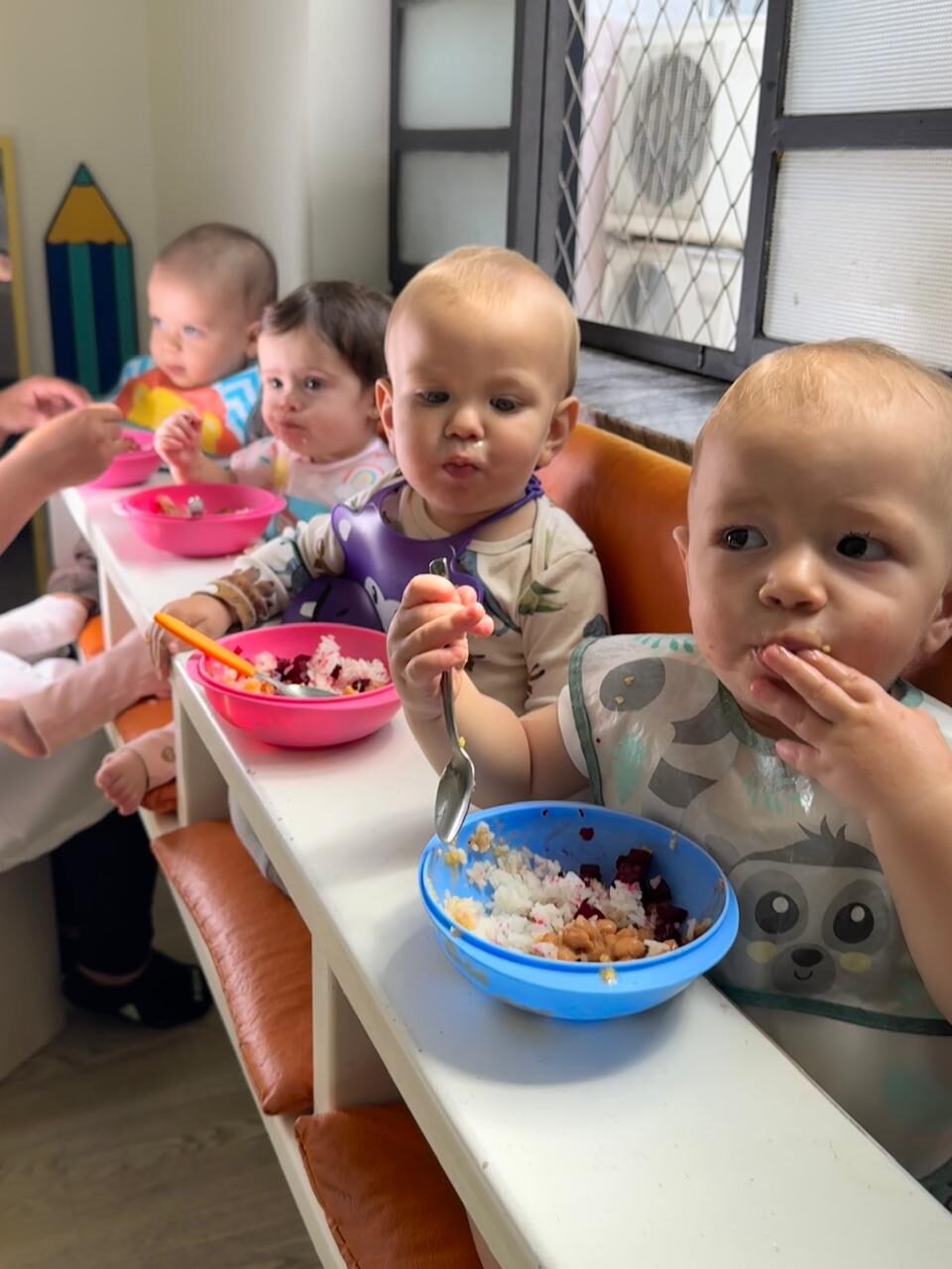 Bebês sentados lado a lado em cadeiras de alimentação, comendo arroz, grãos e beterraba em tigelas coloridas
