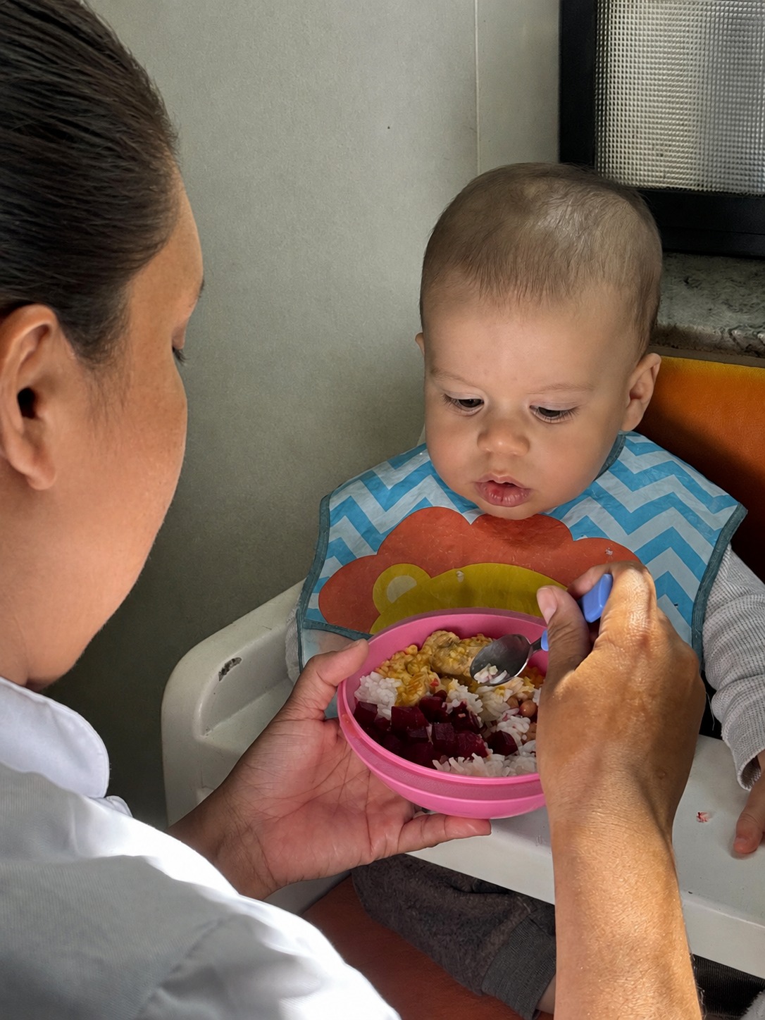 Educadora oferecendo o almoço, com colher, a um bebê sentado na cadeira de alimentação